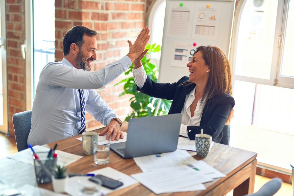 man in white dress shirt sitting beside woman in black long sleeve shirt, giving her a high-five as a business coach.